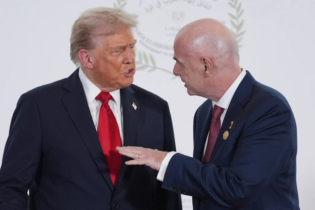 Dwóch mężczyzn w garniturach prowadzi rozmowę, jeden z nich wykonuje gest ręką, a w tle widoczny jest rozmyty herb lub emblemat.