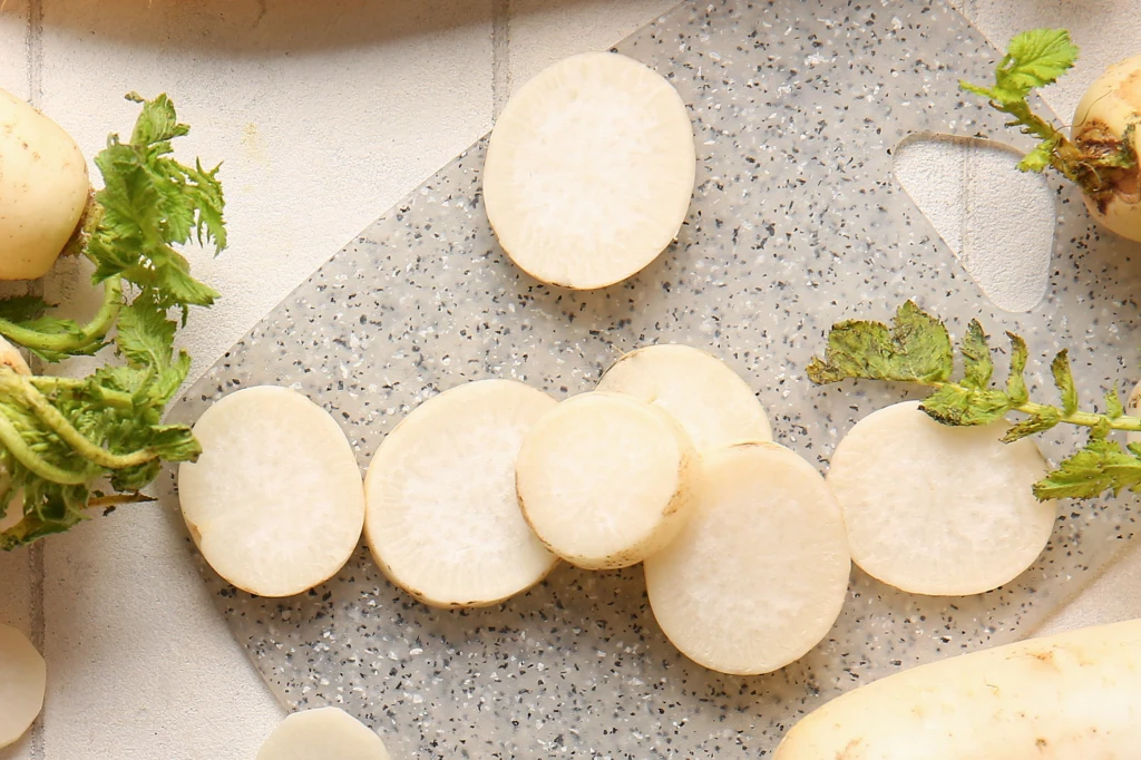 Plasterki białej rzodkwi ułożone na szarej desce do krojenia, wokół widoczne zielone liście i fragmenty reszty warzyw.