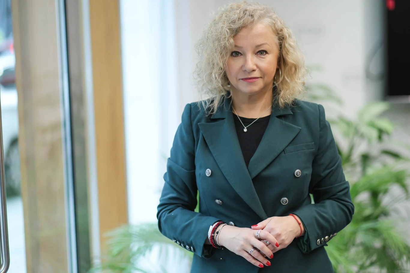 Sekretarz stanu w Kancelarii Prezesa Rady Ministrów Katarzyna Kotula Kobieta o blond, kręconych włosach w zielonej marynarce stoi w jasnym, nowoczesnym wnętrzu z roślinami w tle, patrząc prosto w obiektyw.
