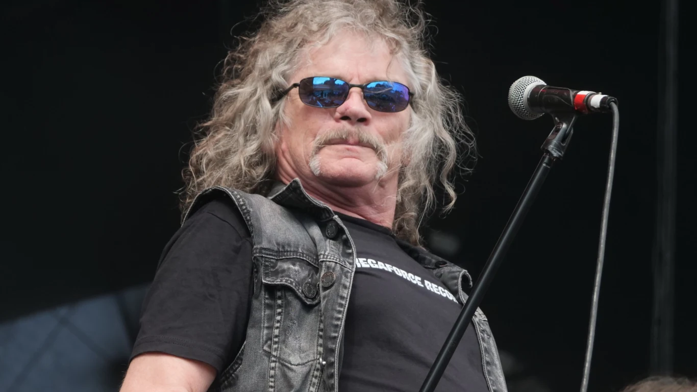 Overkill zagrają na Mystic Festival 2026! Mężczyzna o długich, kręconych, siwych włosach w okularach przeciwsłonecznych i czarnej koszulce stoi na scenie przy mikrofonie, ubrany w kamizelkę jeansową, patrzy w kierunku publiczności.