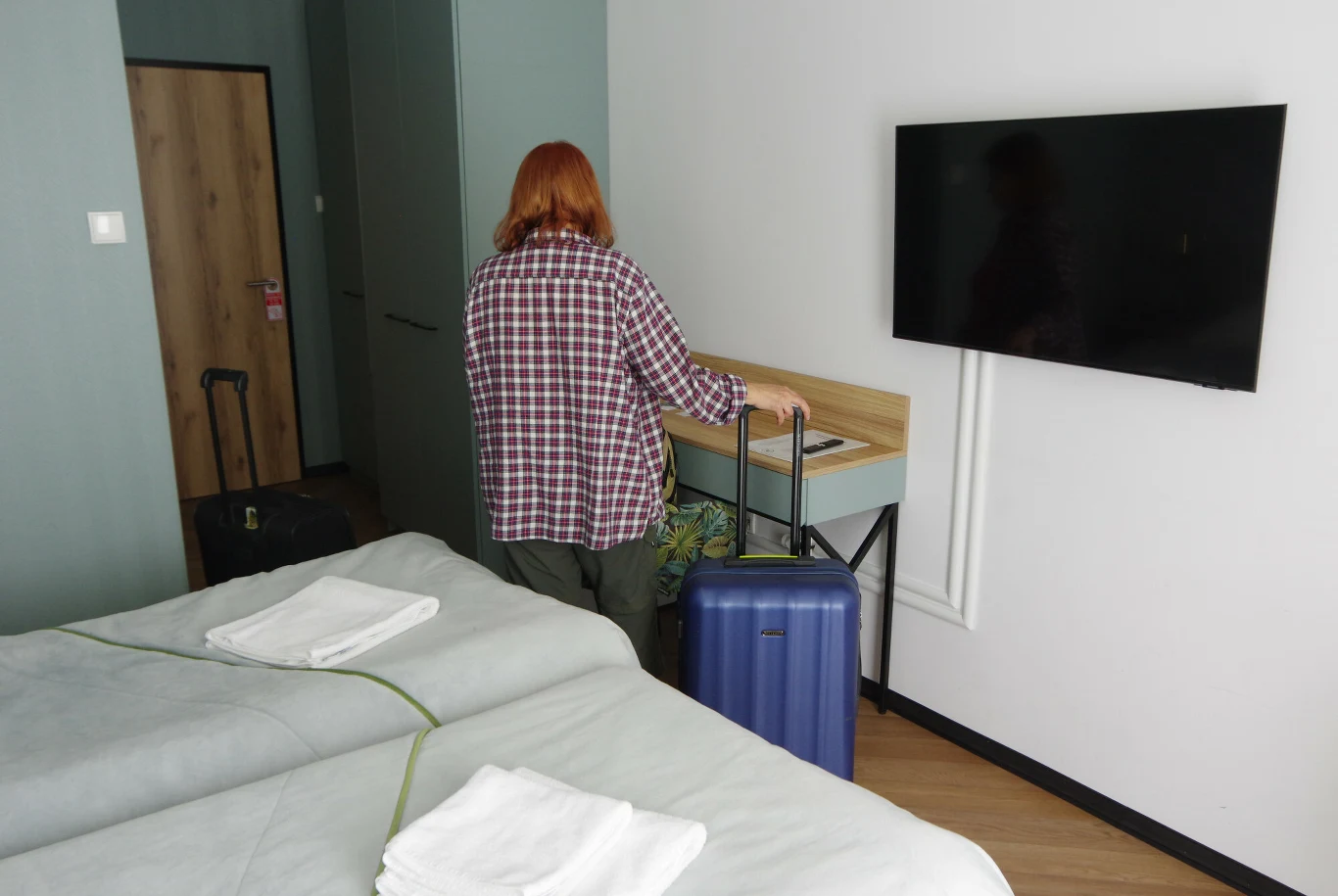 Zmiany w sanatoriach. Co czeka kuracjuszy? Osoba z długimi rudymi włosami w koszuli w kratę stoi przy walizce w hotelowym pokoju; w pomieszczeniu znajduje się telewizor wiszący na ścianie, łóżko z pościelą i ręcznikami oraz walizka i torba podróżna.