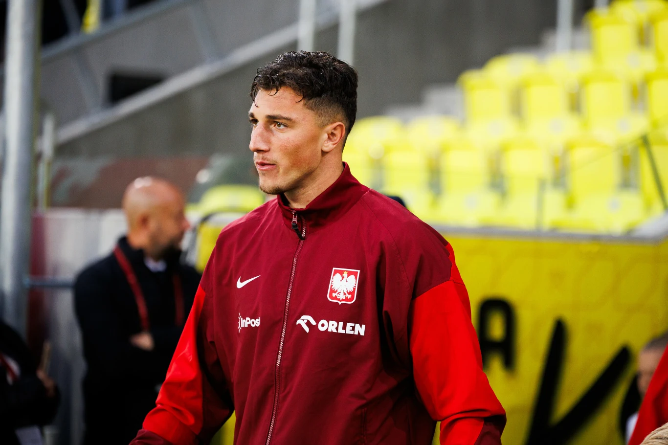 Młody mężczyzna w czerwonej sportowej kurtce z godłem Polski i logotypem Orlen stoi na tle żółtych, stadionowych siedzisk, w tle widoczna rozmyta postać oraz fragment konstrukcji stadionu.