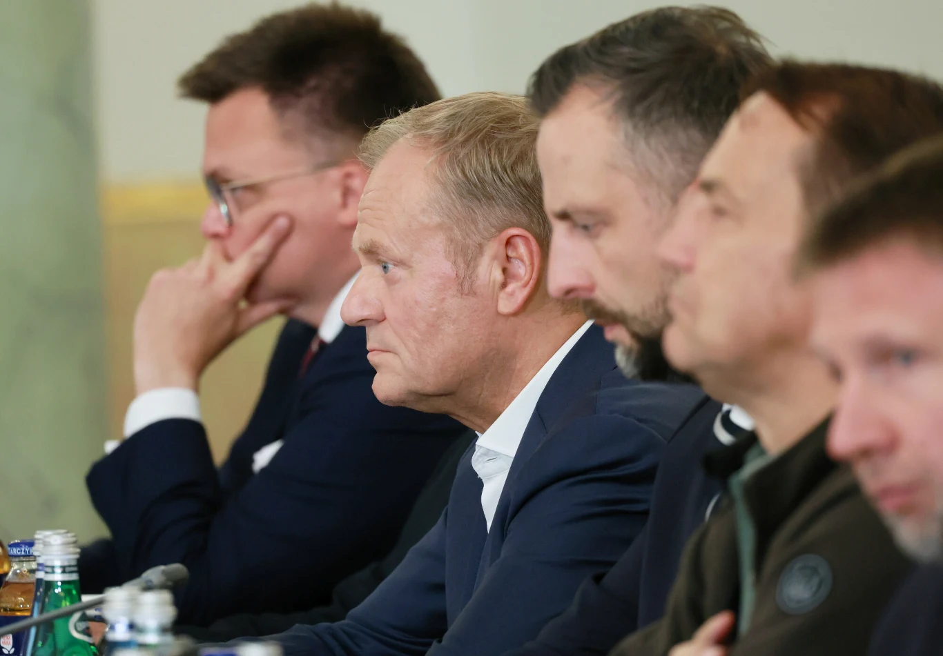 Jaka przyszłość czeka Koalicję 15 Października? Grupa mężczyzn siedzących przy stole podczas oficjalnego spotkania, skupione twarze, w tle butelki z wodą i mikrofony sugerujące przebieg rozmów.