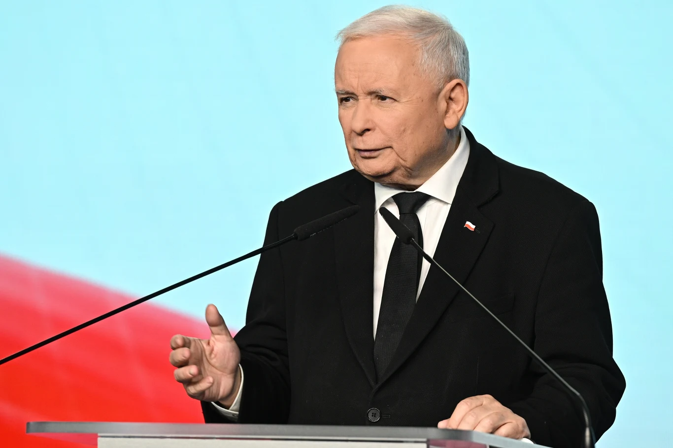 Jarosław Kaczyński Mężczyzna w ciemnym garniturze, z przypiętą flagą Polski na klapie, stoi za mównicą i przemawia z poważnym wyrazem twarzy na tle rozmytego biało-czerwonego tła.