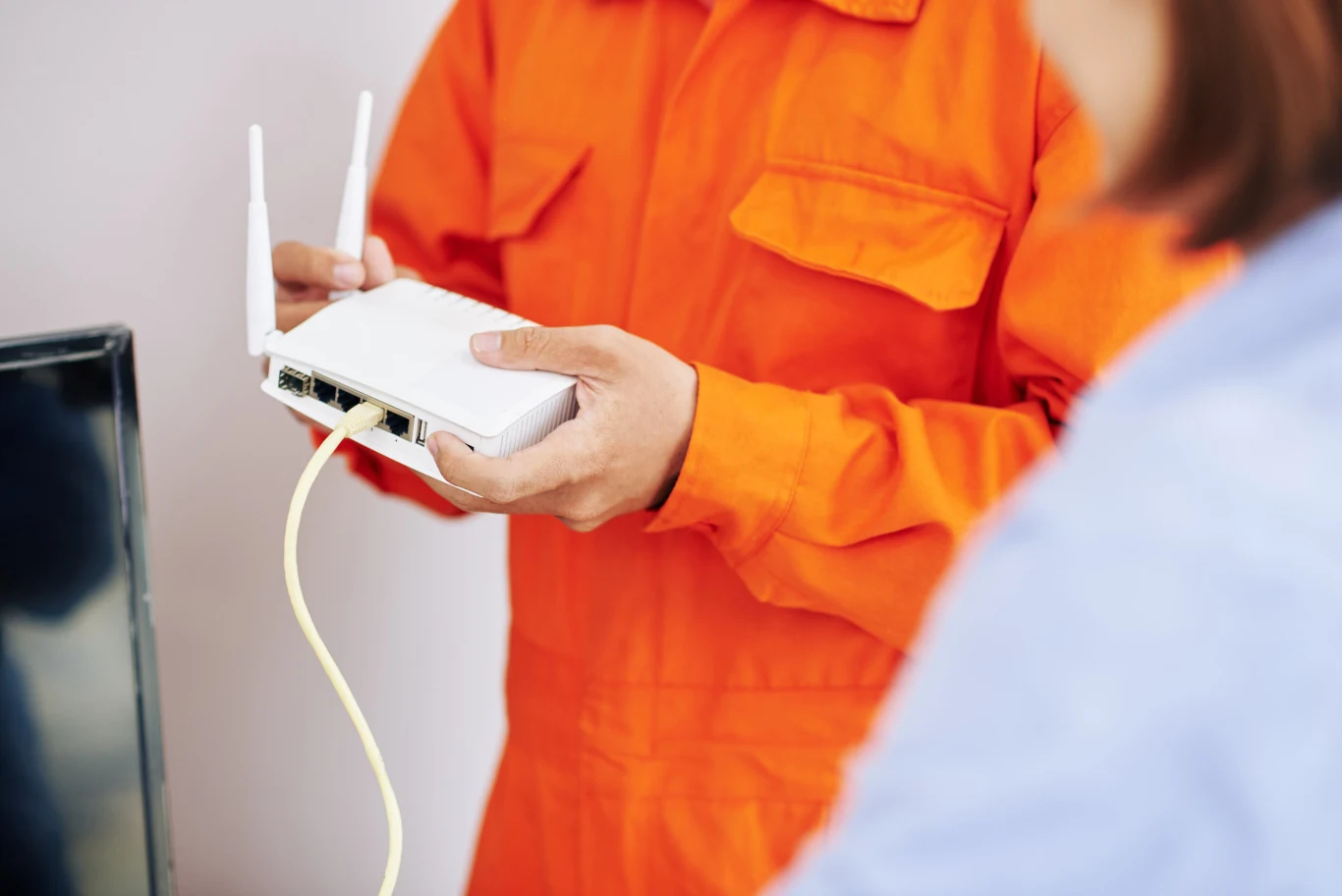 Osoba w pomarańczowym uniformie technicznym trzyma biały router Wi-Fi z podłączonym żółtym kablem sieciowym, obok widoczna sylwetka drugiej osoby.