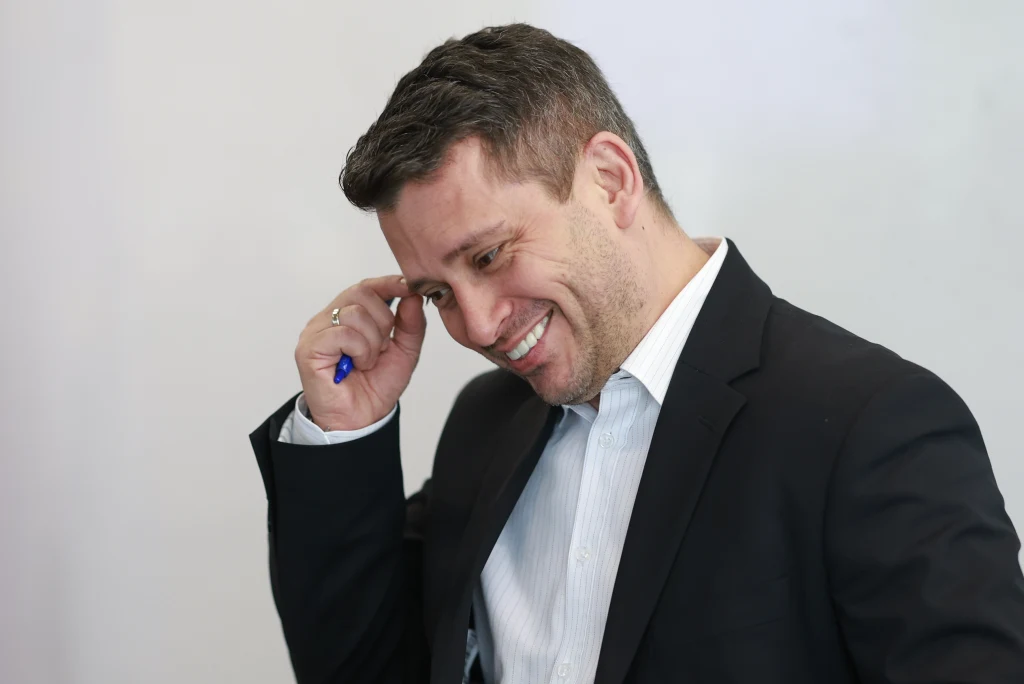 Paweł Śliz Mężczyzna w garniturze z uśmiechem na twarzy trzyma długopis przy głowie, sprawiając wrażenie zamyślonego lub rozbawionego.