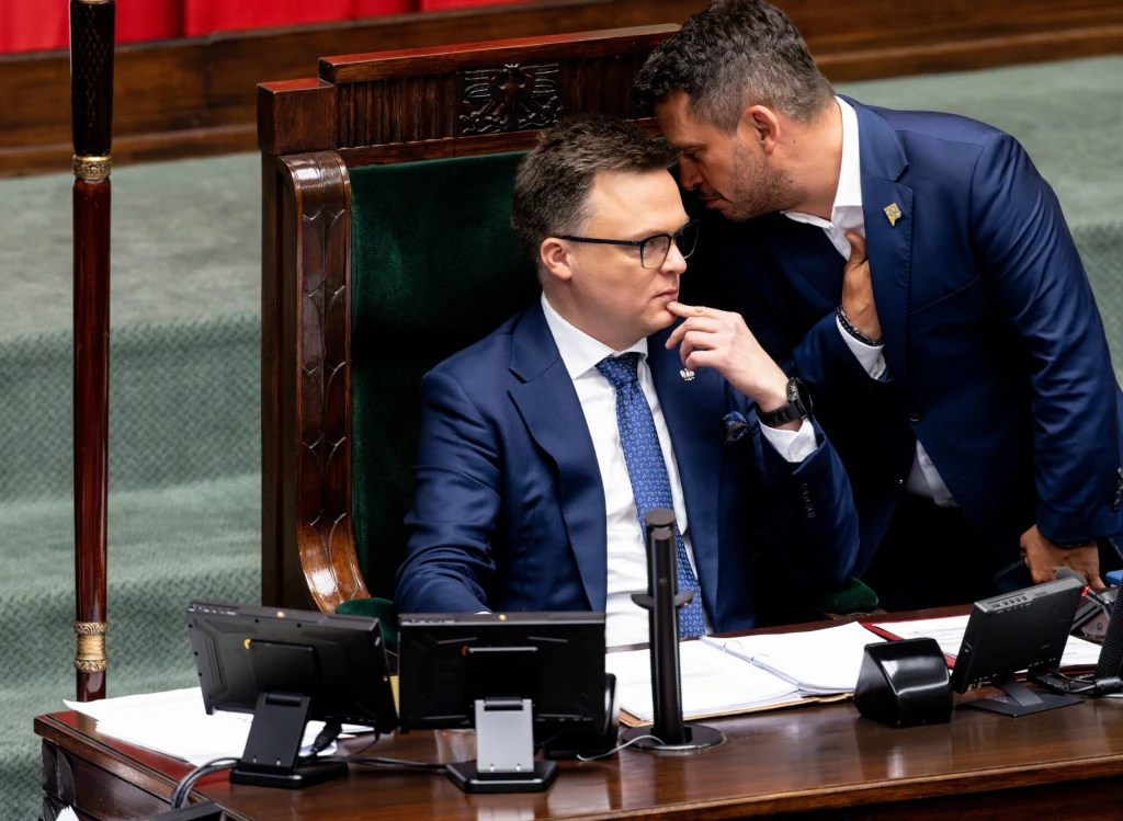 Paweł Śliz podczas narady z Szymonem Hołownią Dwóch mężczyzn w garniturach, jeden siedzi w przewodniczącym fotelu przy biurku z mikrofonem i dokumentami, drugi nachyla się i szepcze mu coś do ucha, scena rozgrywa się w oficjalnej sali o zielonym dywanie