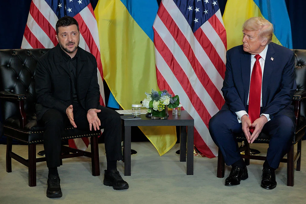 Wołodymyr Zełenski i Donald Trump Dwóch mężczyzn siedzi naprzeciw siebie na czarnych fotelach, pomiędzy nimi stoi stolik z kwiatami i dokumentami, w tle flagi Stanów Zjednoczonych i Ukrainy.