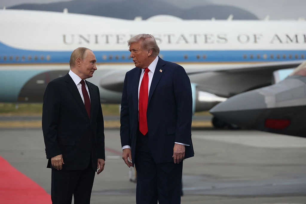 Donald Trump i Władimir Putin Dwóch mężczyzn w garniturach prowadzi rozmowę na płycie lotniska, w tle widoczny samolot rządowy z napisem 'United States of America' i zaparkowany samochód.