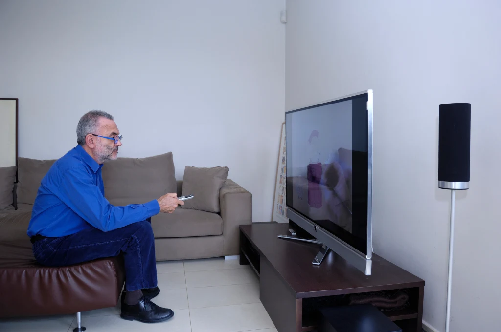 Mężczyzna siedzi na fotelu z pilotem w ręce i włącza telewizor 