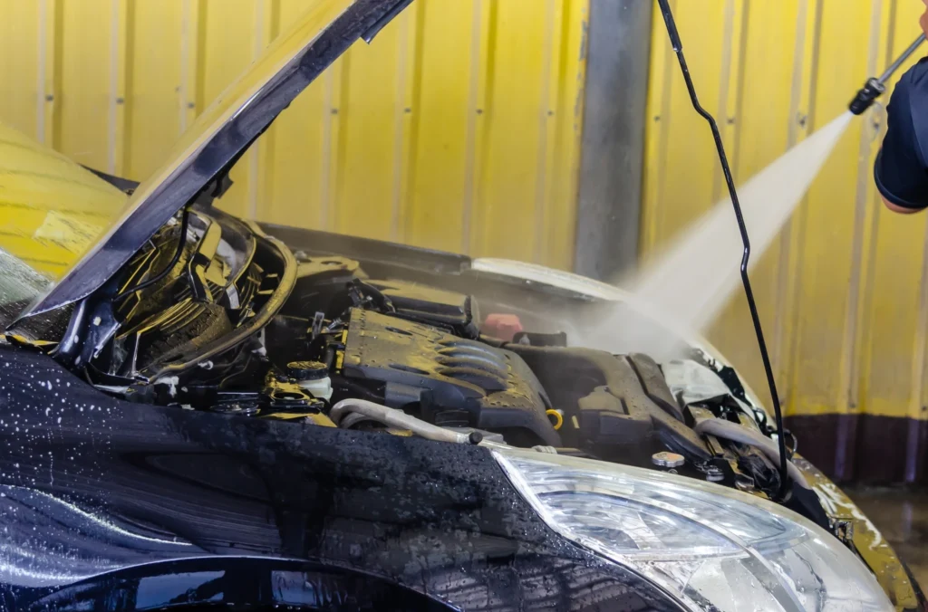 Jak myć silnik w samochodzie? Mycie komory silnika czarnego samochodu ciśnieniową myjką ręczną przy żółtej, metalowej ścianie.