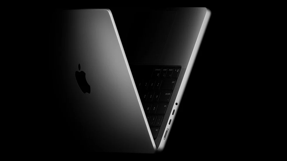 MacBook Pro z czipem Apple M5. Srebrny laptop z charakterystycznym logo Apple, częściowo otwarty, ukazujący porty boczne oraz fragment klawiatury, sfotografowany na czarnym tle.