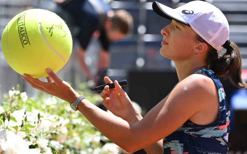 Młoda kobieta w sportowym stroju i czapce z daszkiem trzyma dużą piłkę tenisową oraz marker, przygotowując się do złożenia autografu podczas wydarzenia tenisowego.