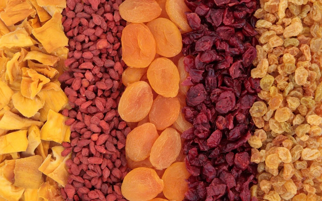 Jak szybko wysuszyć owoce w domu? Kolorowe pasy suszonych owoców ułożone w równoległe rzędy, od lewej żółte mango, czerwone jagody goji, pomarańczowe morele, ciemnoczerwone żurawiny oraz jasnożółte rodzynki.