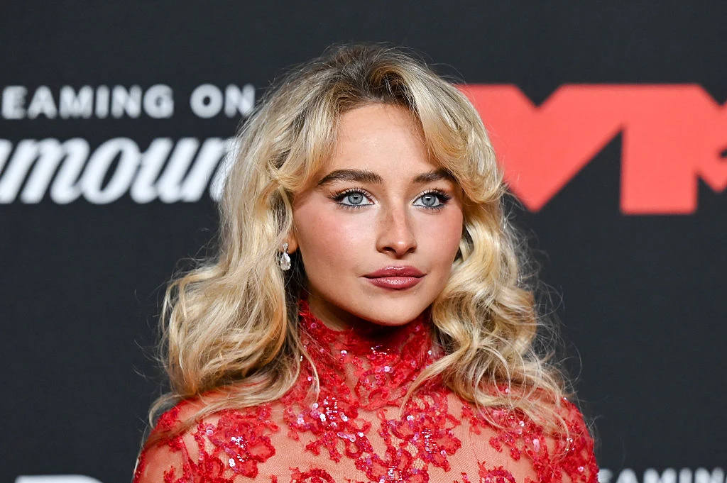 Sabrina Carpenter Kobieta o jasnych włosach i niebieskich oczach w czerwonej, koronkowej sukni, stojąca na tle ścianki z logotypami i napisami.