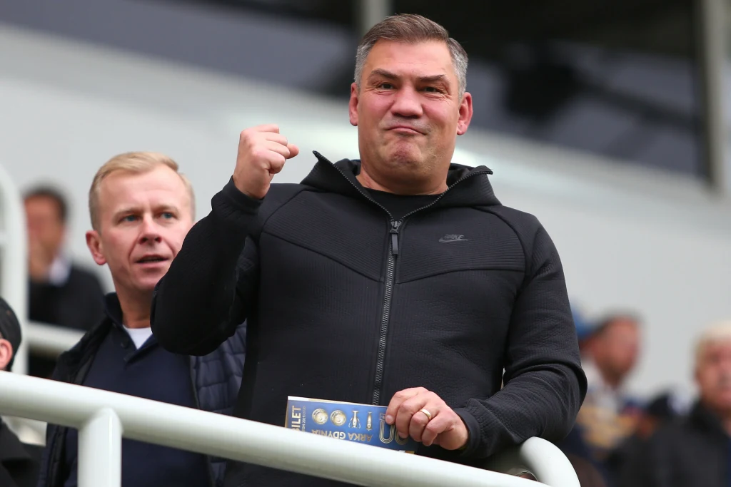 Mężczyzna w czarnej bluzie okazuje radość i zaciska pięść, stojąc przy barierce otoczony innymi osobami na trybunach stadionu.