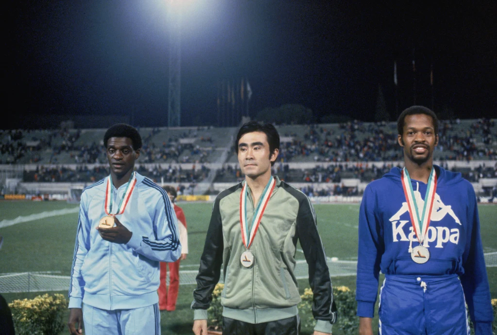 Joao Carlos de Oliveira (pierwszy od lewej) Trzech sportowców stoi na podium podczas ceremonii wręczenia medali na stadionie, wszyscy mają medale na szyjach i sportowe stroje, w tle widoczna jest publiczność.