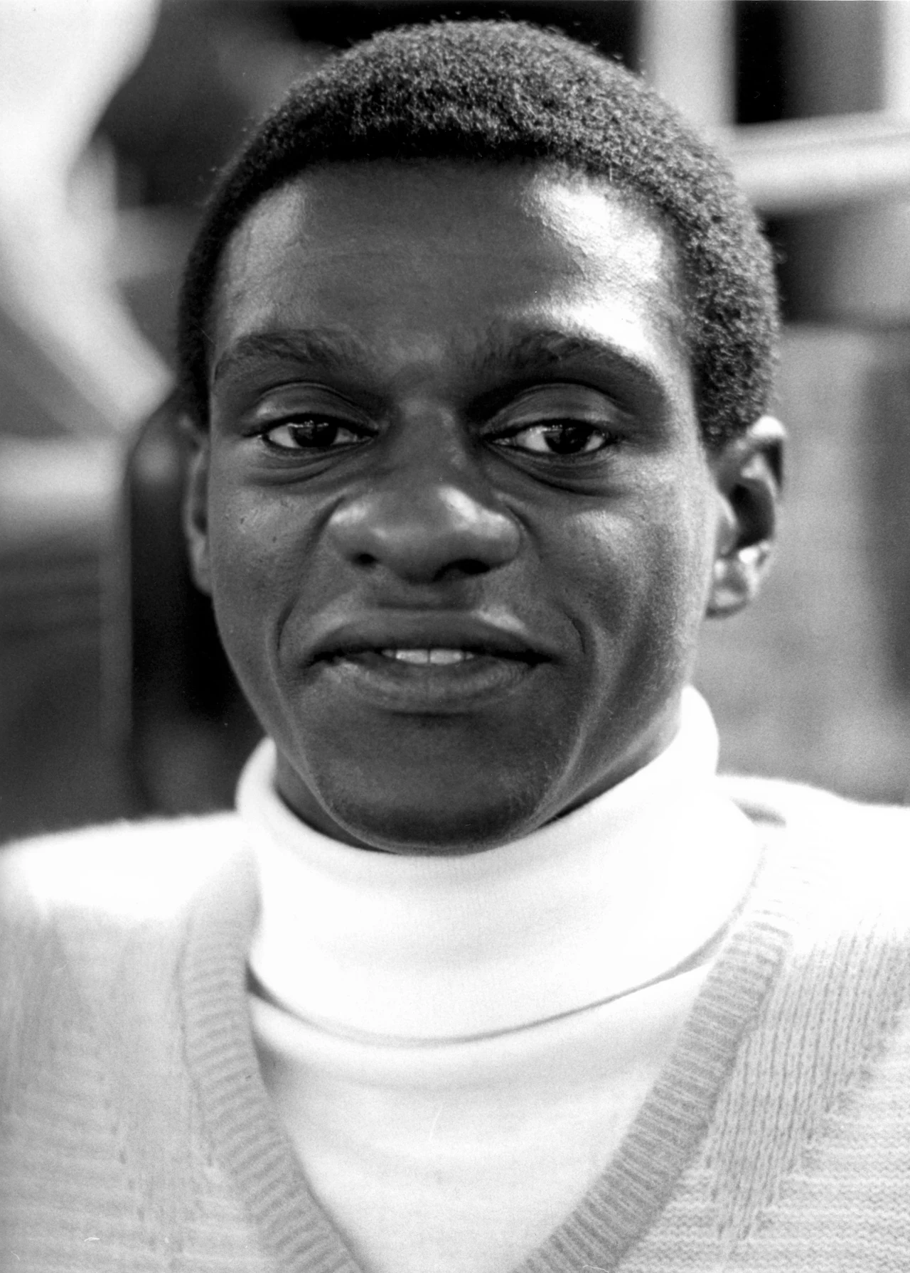 Joao Carlos de Oliveira Uśmiechnięty mężczyzna o krótkich, kręconych włosach, ubrany w jasny sweter z golfem, znajduje się w pomieszczeniu, w tle widoczne rozmyte elementy wnętrza.