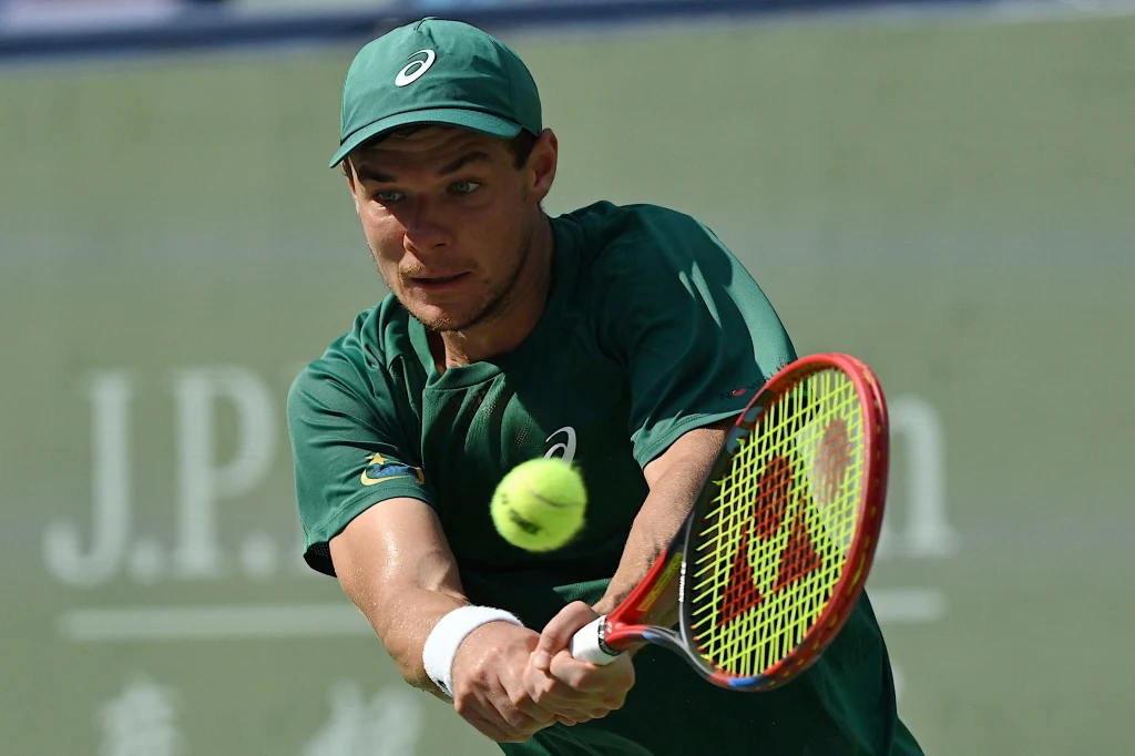 Kamil Majchrzak Mężczyzna w zielonym stroju sportowym podczas dynamicznego uderzenia rakietą do tenisa, zaawansowany moment gry z piłką tuż przed rakietą, tło z logo sponsora.
