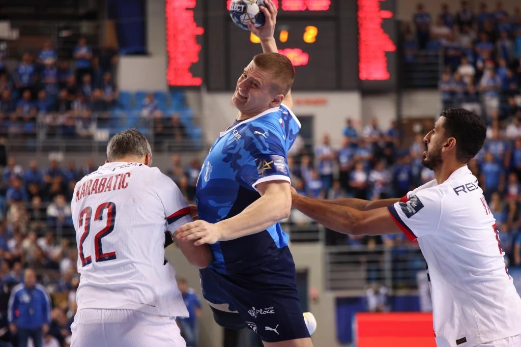 Dawid Dawydzik Dynamiczna akcja podczas meczu piłki ręcznej, zawodnik w niebieskim stroju wykonuje rzut, a dwóch rywali w białych koszulkach próbuje go zablokować, w tle widownia oraz tablica wyników.