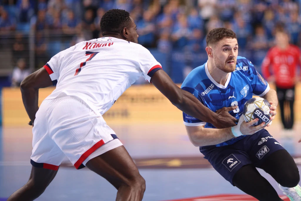 Mitja Janc Dwóch zawodników podczas meczu piłki ręcznej, jeden ubrany w niebieski strój próbuje przedrzeć się z piłką obok drugiego gracza w białym stroju, znajdujących się na hali sportowej przy licznie zgromadzonej publiczności.