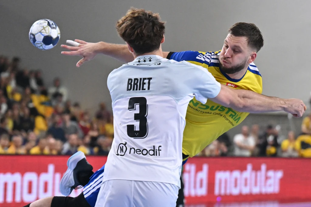 Dynamiczny moment meczu piłki ręcznej, dwóch zawodników w trakcie walki o piłkę, zawodnik w białej koszulce z numerem 3 blokuje atakującego gracza w żółto-niebieskim stroju.