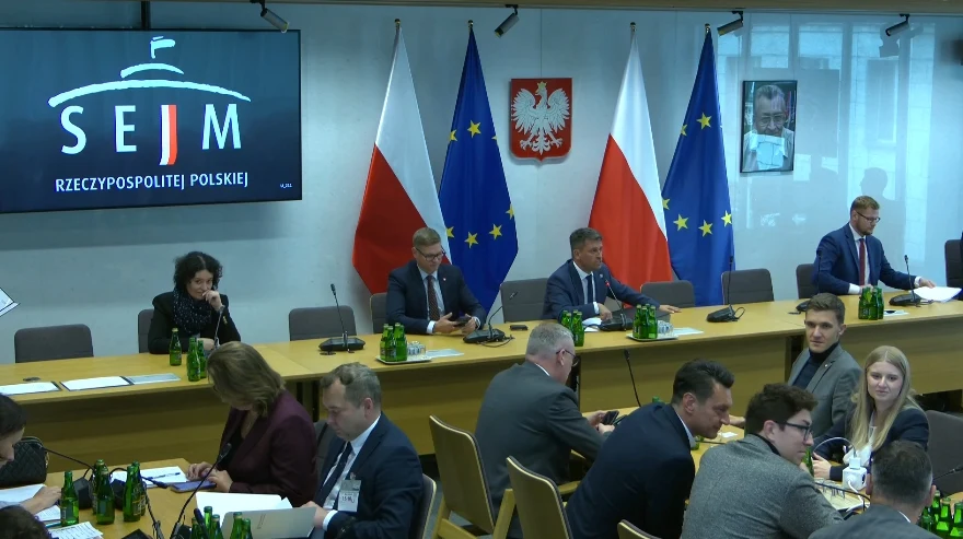 Incydent na posiedzeniu sejmowej komisji sprawiedliwości Oficjalne spotkanie w sali konferencyjnej polskiego Sejmu, przy stole siedzą politycy oraz urzędnicy, na ścianie widoczne flagi Polski i Unii Europejskiej, godło oraz ekran z logo Sejmu, w tle portret Jana Pawła II.