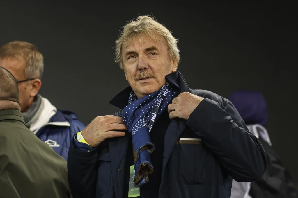 Starszy mężczyzna poprawia niebieski szalik w białe wzory, nosi ciemną kurtkę i stoi na tle ciemnego, rozmytego tła, obok niego widoczny fragment innej osoby.