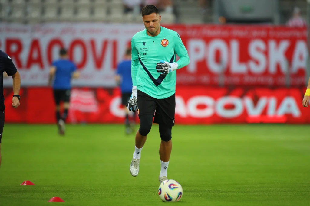 Bramkarz w zielonej koszulce podczas treningu piłkarskiego, żonglujący piłką na murawie, w tle rozmazane postacie i transparent z napisem Polska