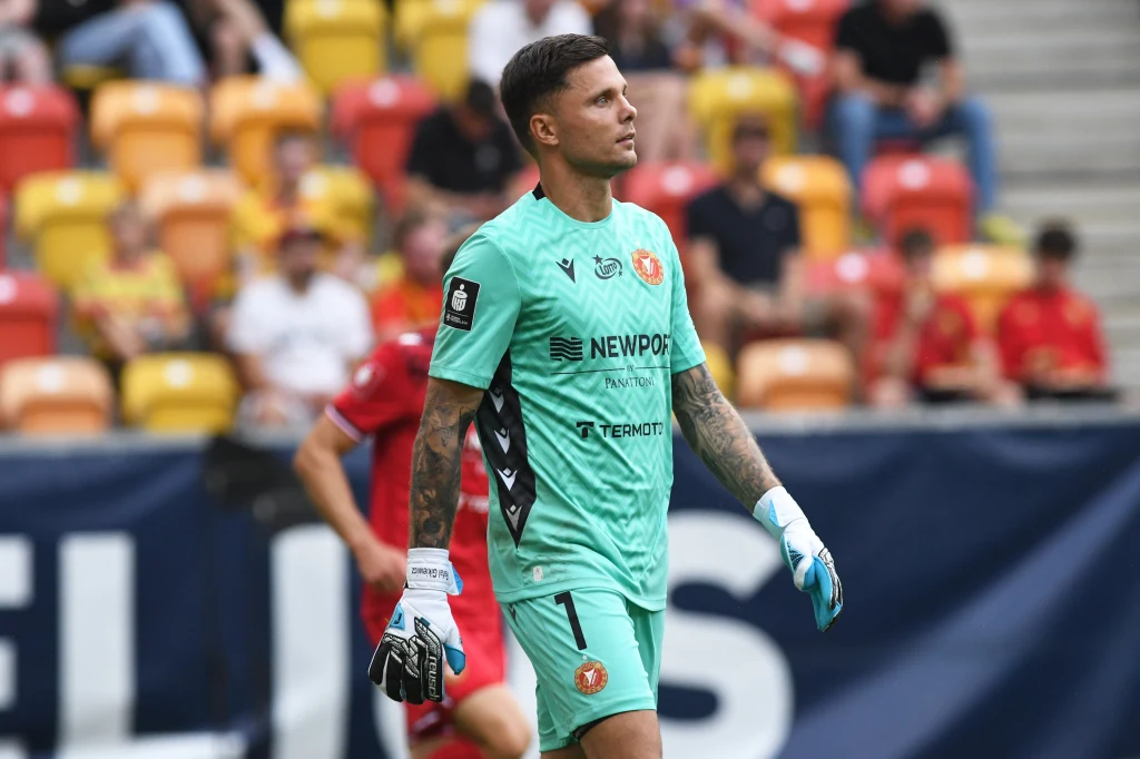 Bramkarz w jasnozielonym stroju piłkarskim stoi na boisku podczas meczu, na tle trybun wypełnionych kibicami ubraną głównie w czerwone i żółte stroje