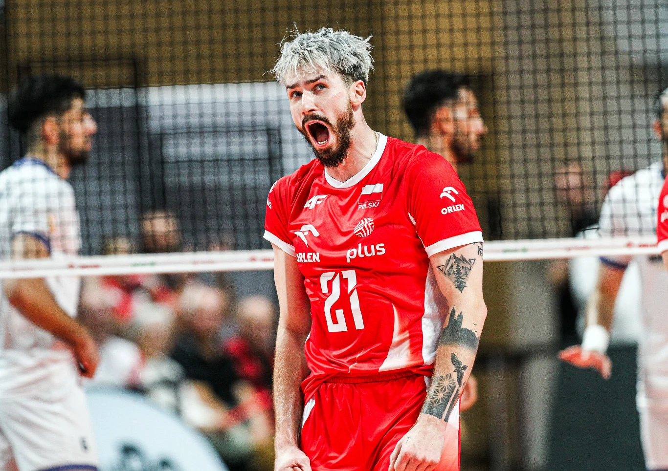 Tomasz Fornal w reprezentacji Polski Zawodnik siatkarski w czerwonym stroju, z widocznym numerem 21, wyraża silne emocje na boisku, stojąc przy siatce, w tle inni gracze oraz rozmyta publiczność.