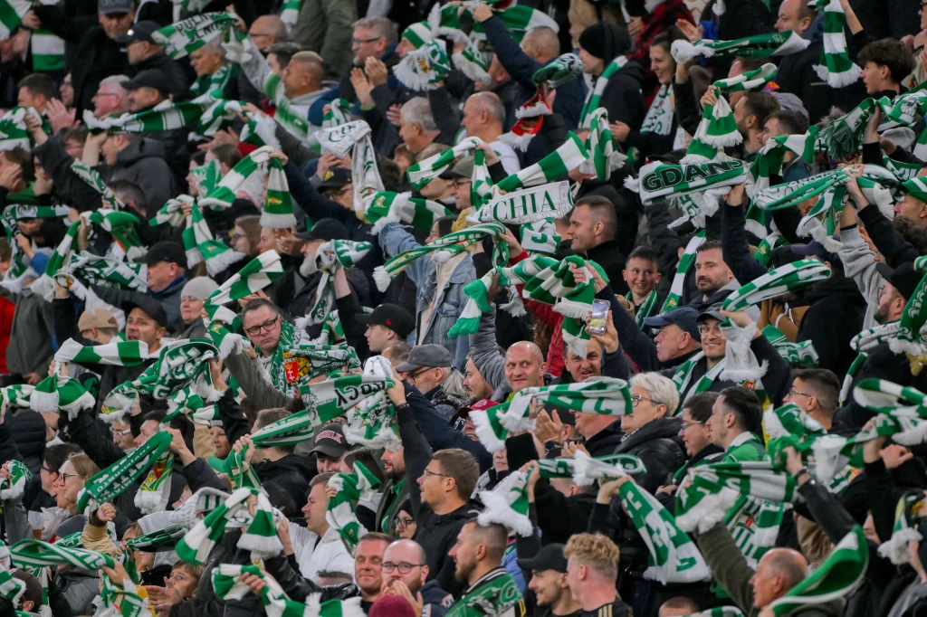 Lechia Gdańsk Tłum kibiców na trybunach stadionu, większość osób trzyma zielono-białe szaliki nad głową, na twarzach widać emocje, a całość tworzy dynamiczną atmosferę sportowego dopingu.