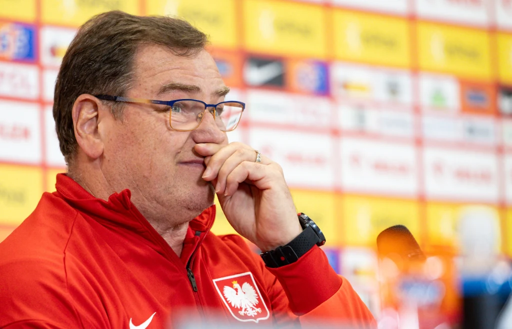 Mężczyzna w okularach ubrany w czerwony dres z godłem Polski, siedzi przy stole konferencyjnym na tle ściany z logotypami sponsorów i partnerów, wydaje się być w trakcie konferencji prasowej.