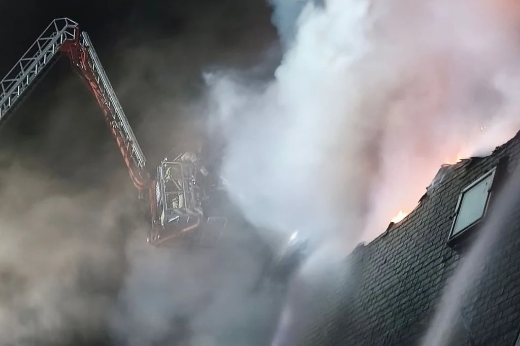 Pożar hotelu w Mniszewie w woj. mazowieckim Gęsty dym i płomienie wydobywające się z dachu budynku, obok którego pracuje podnośnik strażacki z rozłożoną drabiną i koszem.
