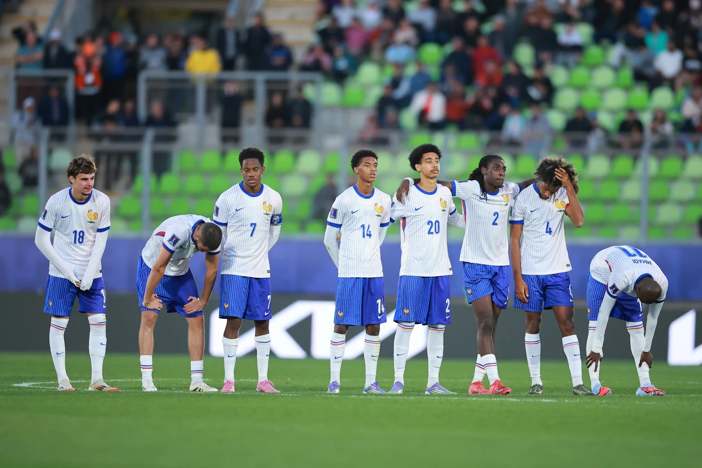 Grupa siedmiu młodych piłkarzy w biało-niebieskich strojach stoi w szeregu na murawie stadionu, kilku z nich wyraźnie okazuje emocje, część pochyla głowy lub trzyma dłonie na kolanach, w tle widoczne są trybuny z kibicami.