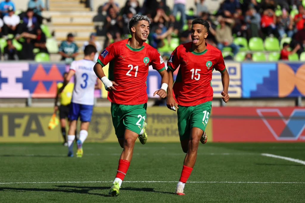 Maroko - Francja, półfinał MŚ U-20 2025 Dwóch młodych piłkarzy w czerwonych koszulkach z numerami 21 i 15 biegnie po murawie stadionu, radośnie celebrując sukces. W tle widoczni są zawodnicy drużyny przeciwnej oraz zielone trybuny wypełnione kibicami.