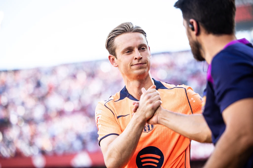 Frenkie de Jong Dwóch mężczyzn podaje sobie dłonie na tle trybun stadionu, jeden ubrany w pomarańczową koszulkę sportową, drugi w granatową koszulkę z mikrofonem.