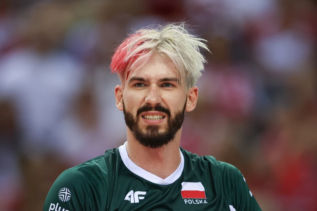Siatkarz w zielonej koszulce reprezentacji Polski z rozjaśnionymi włosami, częściowo zafarbowanymi na czerwono, na tle rozmytej publiczności.