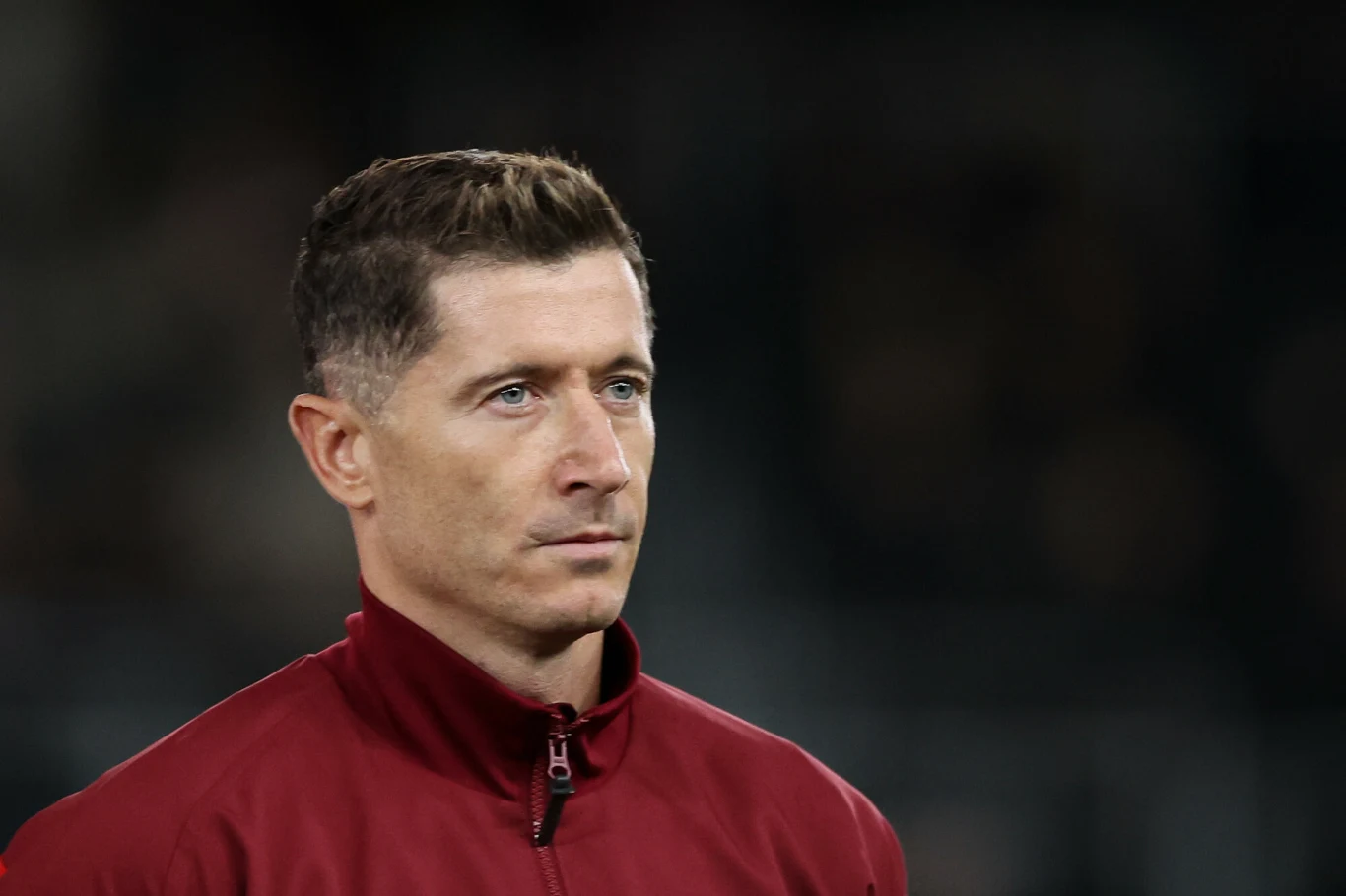 Robert Lewandowski Mężczyzna w bordowej kurtce sportowej na ciemnym, rozmytym tle, lekko zwrócony w bok, z poważnym wyrazem twarzy.