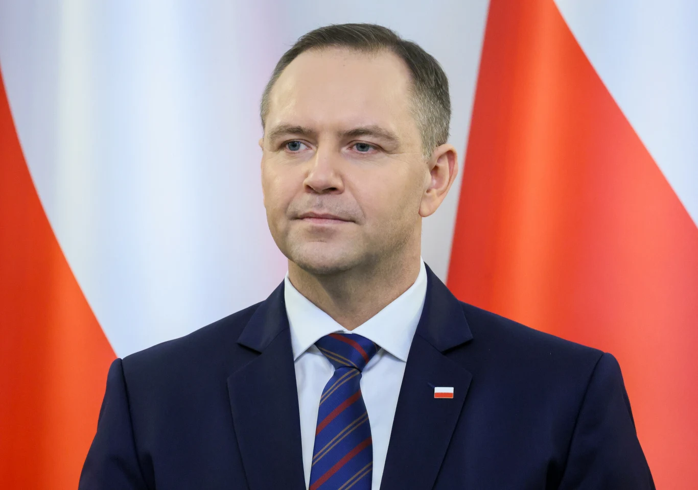 Prezydent Karol Nawrocki Mężczyzna w garniturze i krawacie stoi na tle dużych biało-czerwonych flag, na klapie marynarki widoczna przypinka z flagą Polski