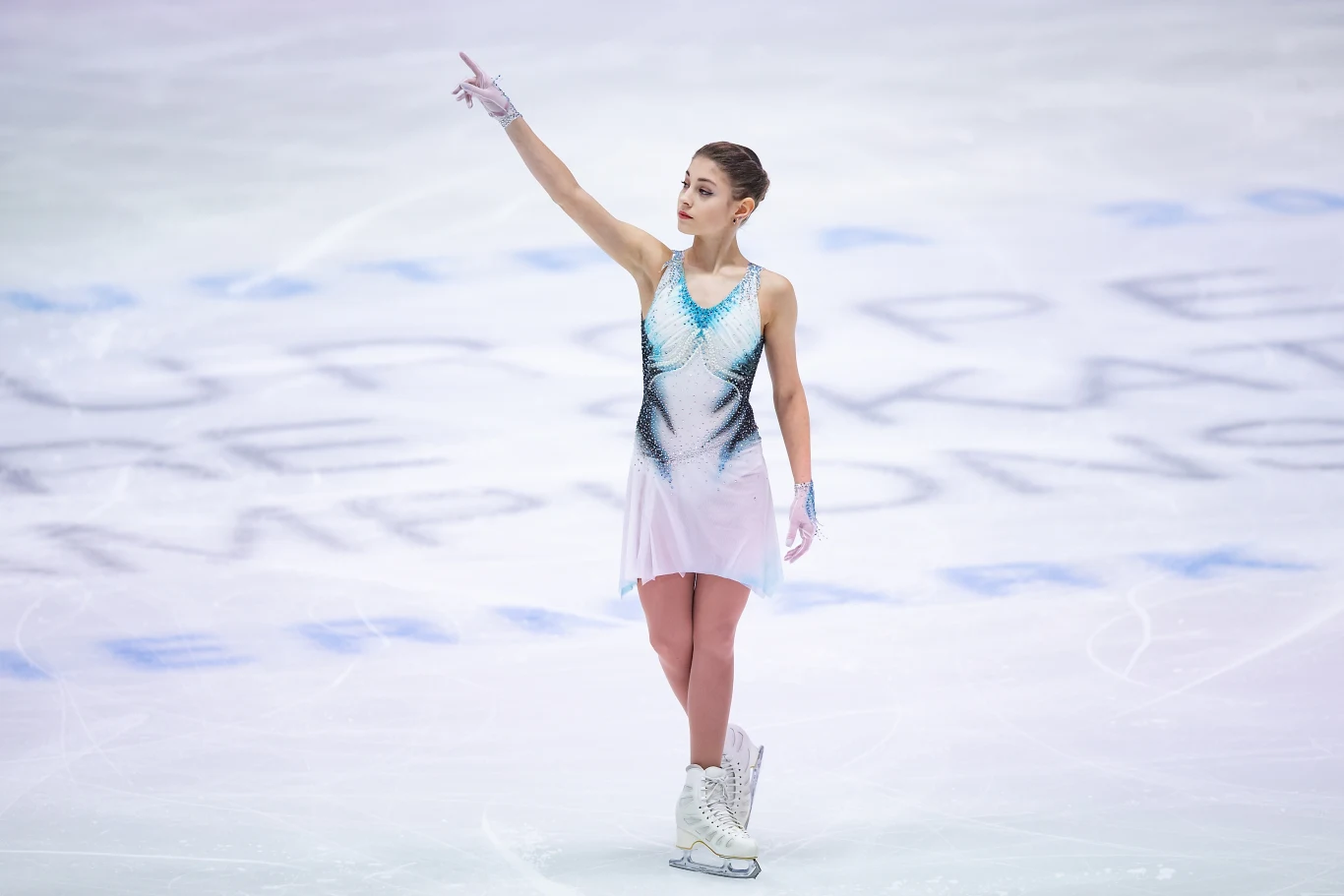 Alona Kostornoj Łyżwiarka figurowa w eleganckim stroju występuje na lodowisku, wykonując choreograficzny układ z uniesioną ręką, w tle widać rozmyte napisy na lodzie.