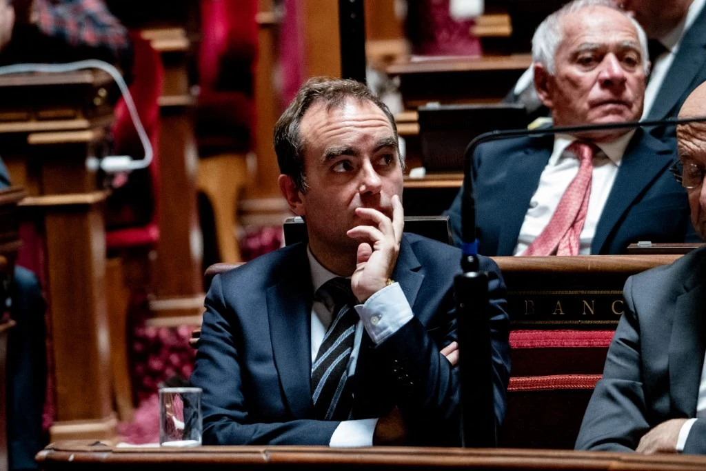 Sebastien Lecornu, premier Francji Mężczyzna w garniturze siedzący na sali obrad, oparty o dłoń, w tle inni uczestnicy posiedzenia również w strojach formalnych, wystrój wnętrza klasyczny z drewnianymi ławami.