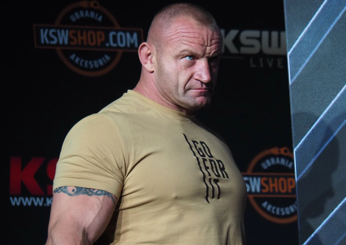 Mariusz Pudzianowski Umięśniony mężczyzna o zdecydowanym wyrazie twarzy w beżowej koszulce z napisem, stojący na tle ciemnych banerów z napisem KSW i logo sklepu internetowego.