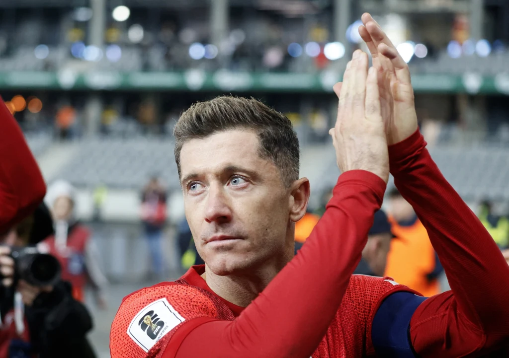 Robert Lewandowski Piłkarz w czerwonym stroju z opaską kapitana na ramieniu składa ręce w geście podziękowania, w tle zamazana publiczność i stadionowe trybuny.