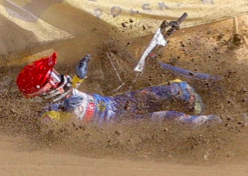 Stal Gorzów na kolejnym zakręcie Zawodnik żużlowy w kasku i pełnym stroju ochronnym upada na torze podczas rywalizacji, wokół niego unosi się piach i fragmenty motocykla, widoczny moment dynamiki upadku