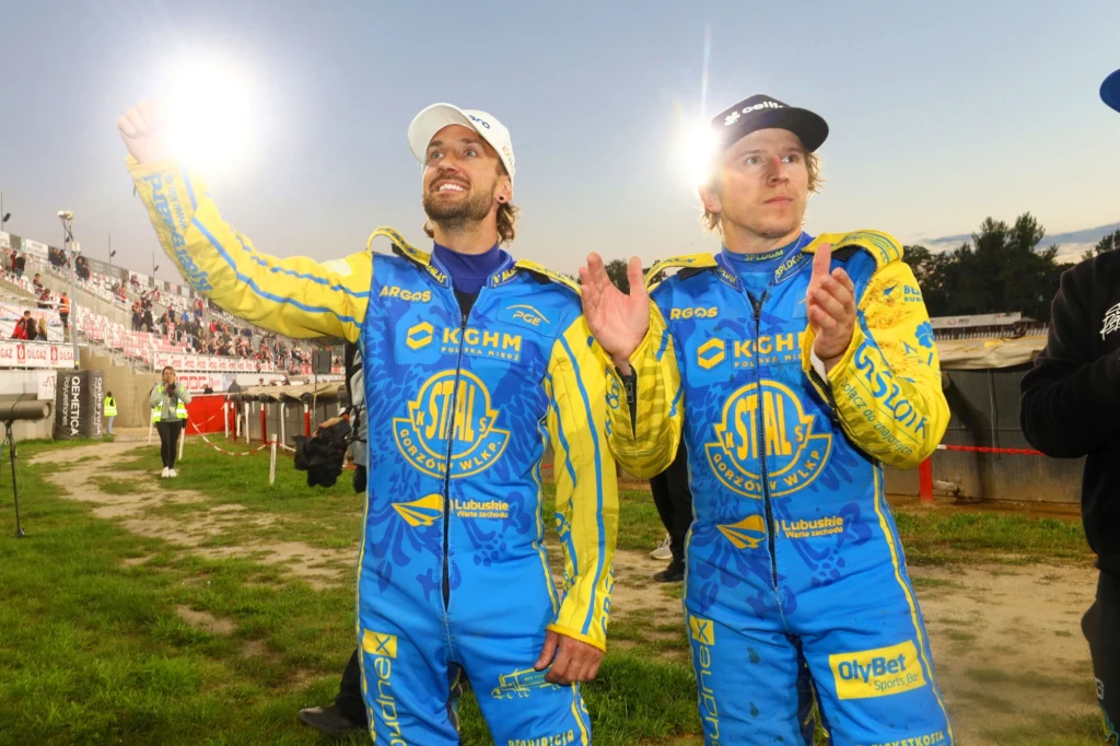 Dwóch sportowców ubranych w żółto-niebieskie kombinezony żużlowe stoi na stadionie, uśmiechają się i machają do kibiców, w tle widoczna część trybun z publicznością oraz otwarta przestrzeń stadionu.