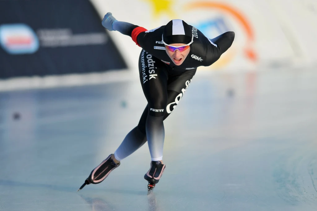 Luiza Złotkowska Łyżwiarz szybki w sportowym kombinezonie w trakcie dynamicznej jazdy na lodowisku, pochylony do przodu w charakterystycznej pozycji aerodynamicznej, w tle rozmazane elementy toru i reklamy.