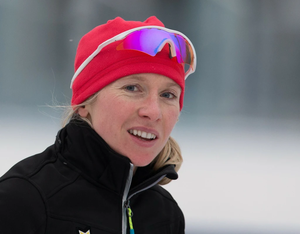 Luiza Złotkowska Kobieta w czerwonej czapce sportowej i okularach przeciwsłonecznych, ubrana w czarną kurtkę, na tle rozmytego zimowego pejzażu.