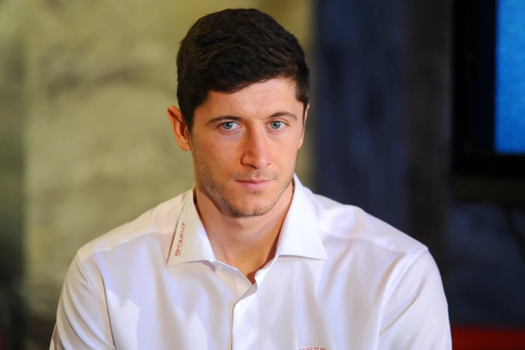 Robert Lewandowski Mężczyzna o krótkich ciemnych włosach w białej koszuli z lekkim zarostem, siedzący na tle rozmytego, neutralnego tła