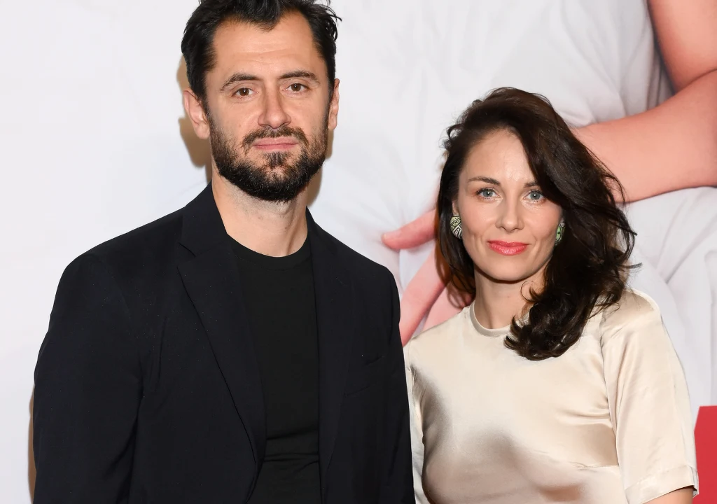 Para elegancko ubranych osób pozuje razem na tle jasnej ściany z elementami graficznymi w tle; kobieta ma na sobie kremową bluzkę, mężczyzna ciemną marynarkę.